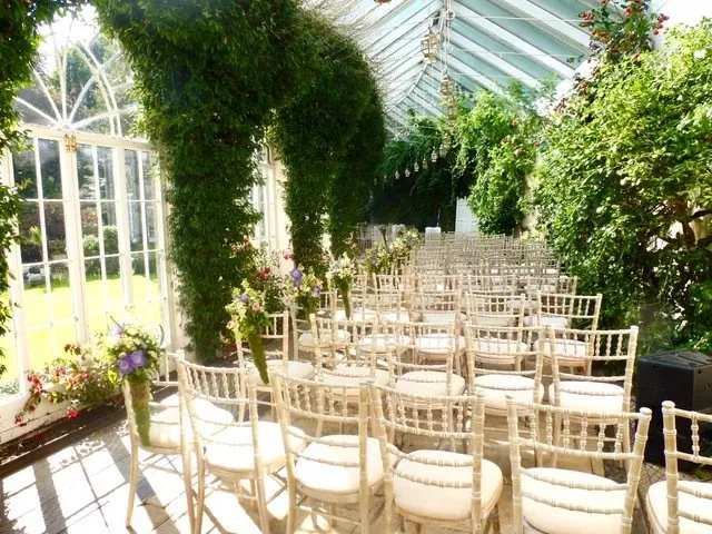 Inside the orangery at Sezincote, set up for a wedding ceremony with chiavari chairs, climbing greenery and arched windows onto the garden